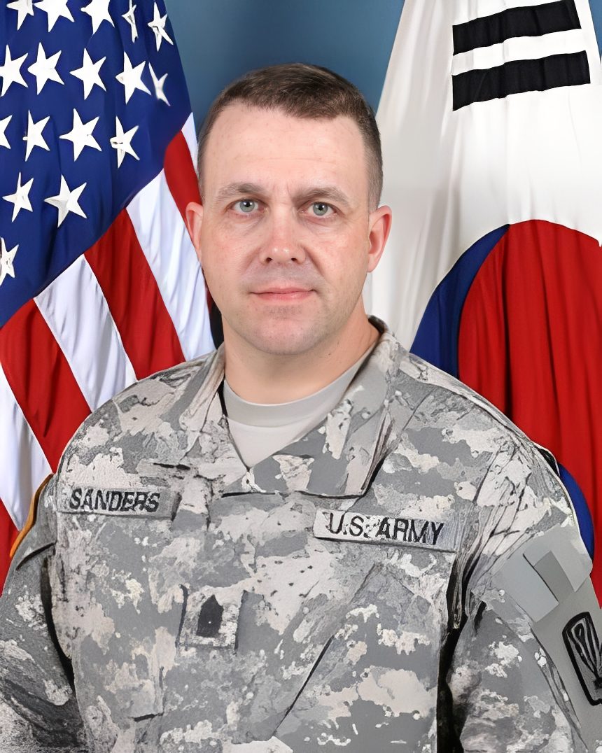 U.S. Army soldier in camouflage, flags behind.